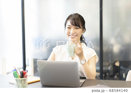 オフィスの席でパソコンの前に座りマグカップを持ち微笑む女性の写真素材