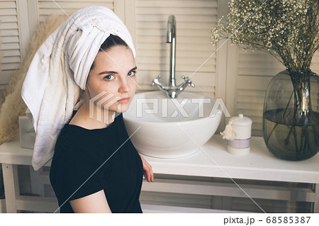 Young beautiful girl with a towel on her head after a shower, in a bathroom with a washbasin and a mirror on the wall. The concept of female morning skin and hair care procedures. 68585387