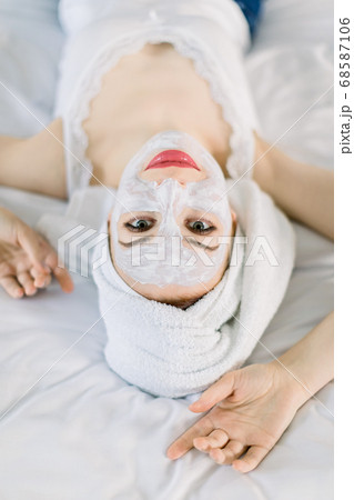 Top close up view of charming smiling Caucasian woman with white clay facial mask and towel on her head, lying on bed and looking at camera, while enjoying skin care treatment Top close up view of charming smiling Caucasian woman with white clay facial mask and towel on her head, lying on bed and looking at camera, while enjoying skin care treatment 68587106