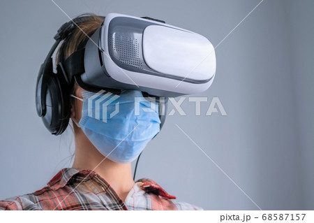 Young woman wearing medical face mask, virtual reality headset against grey wall 68587157