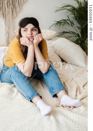 Young beautiful girl student in an orange T-shirt and blue jeans sits on bed with pillows wearing sleep mask. Concept of rest at home, sleep, insomnia, self-isolation, stay home during quarantine. Young beautiful girl student in an orange T-shirt and blue jeans sits on bed with pillows wearing sleep mask. Concept of rest at home, sleep, insomnia, self-isolation, stay home during quarantine. 68600831