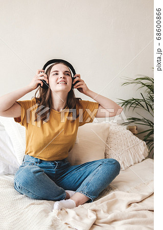 Young beautiful girl student in an orange T-shirt and blue jeans sits on bed with pillows and listens to an audiobook, music, podcast on headphones. Self-education concept at home during quarantine. 68600866