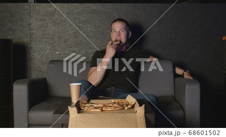 Attractive man eating pizza and watching TV at home using remote control relaxing on couch Attractive man eating pizza and watching TV at home using remote control relaxing on couch 68601502