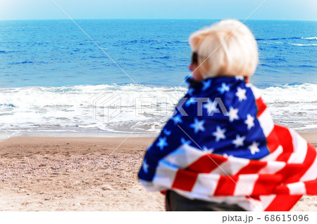 Caucasian blonde woman standing completely wrapped in american flag and looking at sea. Caucasian blonde woman standing completely wrapped in american flag and looking at sea. 68615096