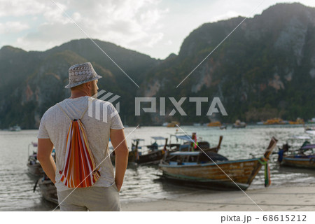 Young male tourist in a hat and with a backpack on a Thai beach on a background of traditional Thai boats. Young male tourist in a hat and with a backpack on a Thai beach on a background of traditional Thai boats. 68615212