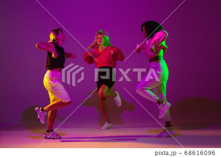 Sportive girls dancing hip-hop in stylish clothes on gradient background at dance hall in neon light Sportive girls dancing hip-hop in stylish clothes on gradient background at dance hall in neon light 68616096