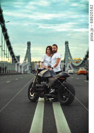 Young sexy woman hugging cute man in stylish black leather jacket, sitting on sports motorcycle on the bridge in the city on sunset and kissing 68620582
