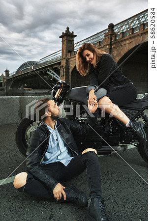 Romantic date on motorbike. Young woman sits on a motorcycle and looks at the man who sits on the ground and holds her hand. Couple in love with sunset under the bridge in the city 68620584
