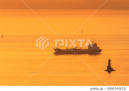 黄金色に輝く夜明けの東京湾で灯台の傍に停泊する貨物船の写真素材