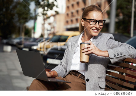 Working from everywhere. Portrait of a happy beautiful business woman drinking coffee and using laptop while sitting on the bench outdoors Working from everywhere. Portrait of a happy beautiful business woman drinking coffee and using laptop while sitting on the bench outdoors 68639637