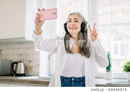 Portrait of positive, happy, smiling senior lady with long gray hair, with headphones, having fun in kitchen at home, listening to music and making photo on cell phone gesturing victory sign Portrait of positive, happy, smiling senior lady with long gray hair, with headphones, having fun in kitchen at home, listening to music and making photo on cell phone gesturing victory sign 68646874