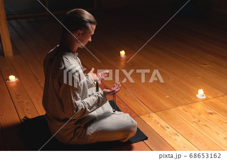 A man in clothes for practice and meditation sits in a lotus pose and holds red rosary to concentrate attention in a wooden room with dim light A man in clothes for practice and meditation sits in a lotus pose and holds red rosary to concentrate attention in a wooden room with dim light 68653162