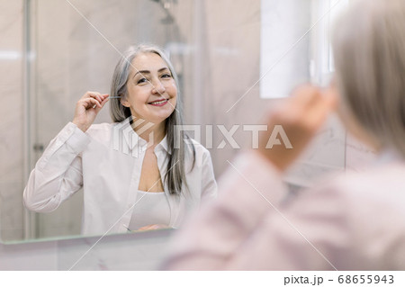 Portrait of pretty senior gray haired woman looking to mirror, cleansing her ears with a cotton swab in modern light bathroom at home. Beauty, hygiene and people concept Portrait of pretty senior gray haired woman looking to mirror, cleansing her ears with a cotton swab in modern light bathroom at home. Beauty, hygiene and people concept 68655943