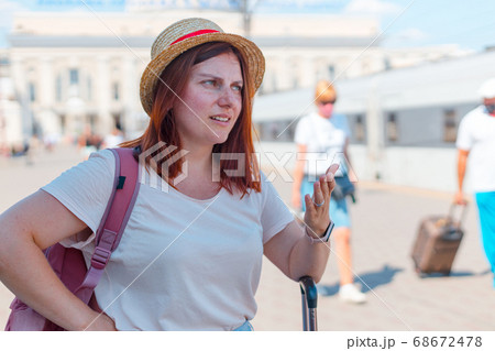 Tired young red-haired girl in a hat with a backpack and suitcase stands at the train station Tired young red-haired girl in a hat with a backpack and suitcase stands at the train station 68672478