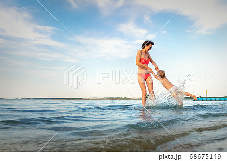 Beautiful young adult caucasian mom enjoy having fun at lake river beach spin little toddler boy son around making water splash. Family travel and vacation with children concept. Outdoor activities 68675149