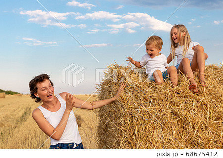 Young adult attractive beautiful mom with little son and daughter enjoy having fun fooling around sitting near golden hay bale on wheat harvested field near farm. Happy children on rural landscape 68675412