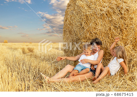 Young adult attractive beautiful mom with little son and daughter enjoy having fun fooling around sitting near golden hay bale on wheat harvested field near farm. Happy children on rural landscape 68675415