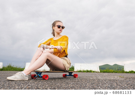 A young attractive girl in a yellow sweater shorts and sunglasses with a tattoo on her arm sits on a longboard behind a suburban asphalt pad 68681133