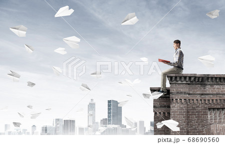 Handsome student guy on roof edge reading book and paper planes fly around Handsome student guy on roof edge reading book and paper planes fly around 68690560