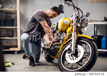 A young sports motorcycle repairman in a garage or workshop is working on a yellow bike. A young sports motorcycle repairman in a garage or workshop is working on a yellow bike. 68703218