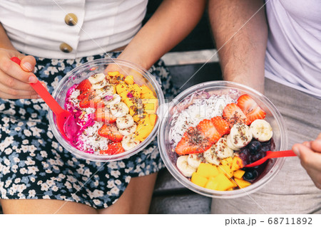 Acai bowl and pitaya dragonfruit smoothie healthy breakfast bowls young friends eating together. Couple man and woman eating sitting outside in park for lunch break. Closeup on food Acai bowl and pitaya dragonfruit smoothie healthy breakfast bowls young friends eating together. Couple man and woman eating sitting outside in park for lunch break. Closeup on food 68711892