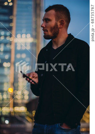Man using smartphone texting. Male standing in the city in the evening outdoors using cell phone and waiting for taxi or uber. Young businessman looking at mobile phone Man using smartphone texting. Male standing in the city in the evening outdoors using cell phone and waiting for taxi or uber. Young businessman looking at mobile phone 68735767