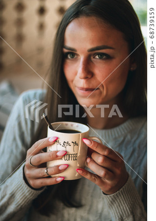 Close up portrait of a beautiful young woman drinking a cup of coffee by a window at home 68739159
