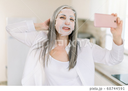 Beautiful retired grau haired lady in white shirt, standing at home kitchen and holding her cell phone, relaxing during beauty procedures with sheet cloth mask and smiling to camera Beautiful retired grau haired lady in white shirt, standing at home kitchen and holding her cell phone, relaxing during beauty procedures with sheet cloth mask and smiling to camera 68744591