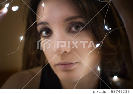 Indoors female portrait of a woman with dreadlocks hairstyle and smoky eyes makeup holding lights garland near her face, femininity and hollidays concepts 68745238