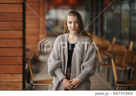 A girl with blond hair and blue eyes in a white jacket and black blouse near the summer cafe stands and holds the phone in her hand. Growth photography 68745969