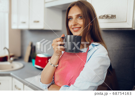 Pretty young woman in red apron enjoying a cup of coffee in her kitchen 68754163