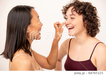 Two young laughing intercultural brunettes having fun during skincare procedure Two young laughing intercultural brunettes having fun during skincare procedure 68765325