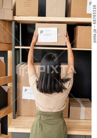 Back view of young female manager with dark long hair taking box from shelf Back view of young female manager with dark long hair taking box from shelf 68765536