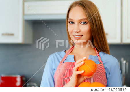 Beautiful caucasian woman in red apron holding ripe oranges while standing in kitchen Beautiful caucasian woman in red apron holding ripe oranges while standing in kitchen 68766612
