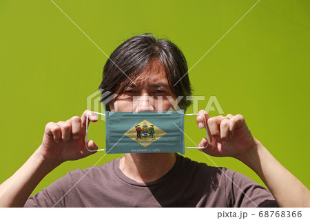 A man and hygienic mask with Delaware flag pattern in his hand and raises it to cover his face. A man and hygienic mask with Delaware flag pattern in his hand and raises it to cover his face. 68768366