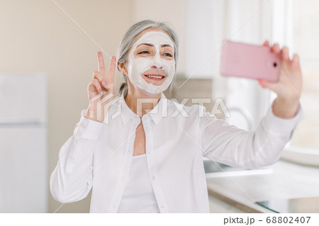 Portrait of positive, happy, smiling senior lady with long gray hair, with mud facial mask, having fun in kitchen at home, making photo on cell phone gesturing victory sign Portrait of positive, happy, smiling senior lady with long gray hair, with mud facial mask, having fun in kitchen at home, making photo on cell phone gesturing victory sign 68802407