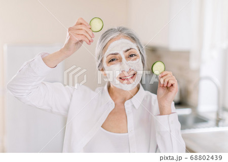 Smiling elderly Caucasian woman with clay mask for face skin and cucumbers slices in hands, posing at light home kitchen, enjoying spa beauty procedures at home. Promo of healthy lifestyle and beauty Smiling elderly Caucasian woman with clay mask for face skin and cucumbers slices in hands, posing at light home kitchen, enjoying spa beauty procedures at home. Promo of healthy lifestyle and beauty 68802439
