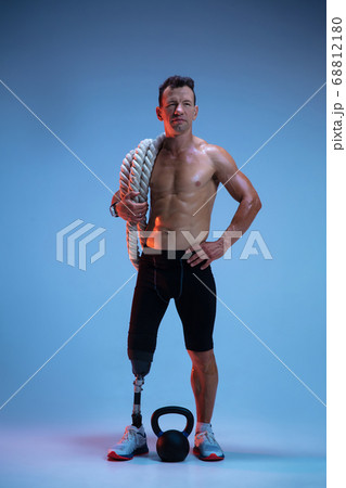 Athlete with disabilities or amputee isolated on blue studio background. Professional male sportsman with leg prosthesis training with weights in neon Athlete with disabilities or amputee isolated on blue studio background. Professional male sportsman with leg prosthesis training with weights in neon 68812180
