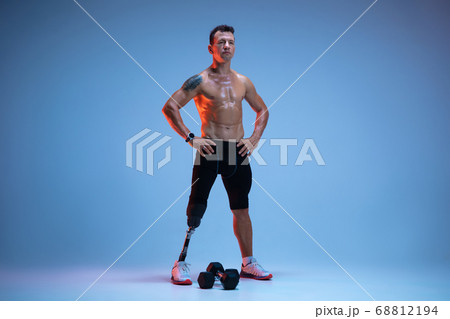 Athlete with disabilities or amputee isolated on blue studio background. Professional male sportsman with leg prosthesis training with weights in neon 68812194
