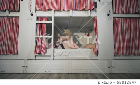 Young attractive woman sitting on the bed in the hostel room and using smartphone Young attractive woman sitting on the bed in the hostel room and using smartphone 68812847