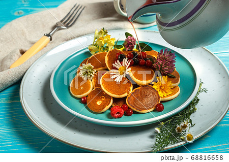 A portion of mini pancakes on a blue plate, decorated with fresh flowers and a jug of pouring syrup. Close up A portion of mini pancakes on a blue plate, decorated with fresh flowers and a jug of pouring syrup. Close up 68816658