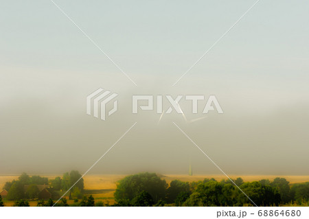 Wind turbines in morning mist. Only part of the turbines are visible in the mist. Yellow straw field an green bushes in the foreground Wind turbines in morning mist. Only part of the turbines are visible in the mist. Yellow straw field an green bushes in the foreground 68864680