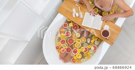 Faceless female in bathtub with slices of oranges, lemon and grapefruits is reading the book. Woman having detox bath treatment. Pedicure tools on wooden tray. 68866924
