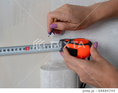 A woman makes measurements on the wall for wallpapering with a measuring ruler. A woman makes measurements on the wall for wallpapering with a measuring ruler. 68884744