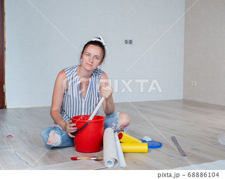 A woman stirs glue in a red bucket for wallpapering in an apartment. A woman stirs glue in a red bucket for wallpapering in an apartment. 68886104