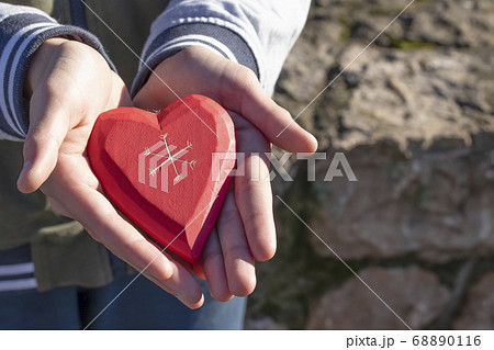 A boy holds and shows a wooden red heart in his hand, being in a park among trees on a sunny Valentine's Day and Mother's Day A boy holds and shows a wooden red heart in his hand, being in a park among trees on a sunny Valentine's Day and Mother's Day 68890116