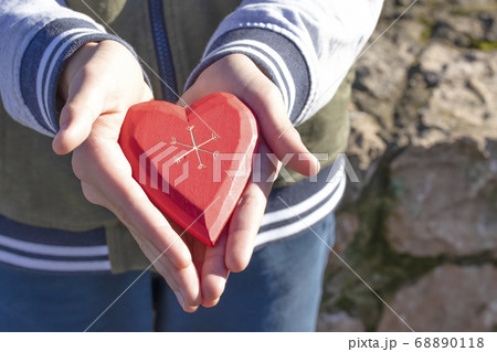 A boy holds and shows a wooden red heart in his hand, being in a park among trees on a sunny Valentine's Day and Mother's Day A boy holds and shows a wooden red heart in his hand, being in a park among trees on a sunny Valentine's Day and Mother's Day 68890118