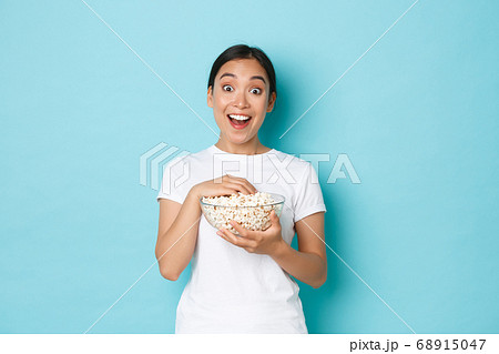 Lifestyle, leisure and emotions concept. Excited and upbeat asian girl in white t-shirt, looking amazed at TV screen, watching movie and eating popcorn, smiling thrilled, light blue background Lifestyle, leisure and emotions concept. Excited and upbeat asian girl in white t-shirt, looking amazed at TV screen, watching movie and eating popcorn, smiling thrilled, light blue background 68915047
