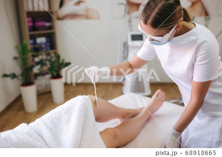 Young woman beautician in mask applies contact gel on the leg of a woman for laser hair removal in medical clinic Young woman beautician in mask applies contact gel on the leg of a woman for laser hair removal in medical clinic 68918665