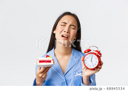 Close-up of displeased asian girl in blue pajama express regret as cant eat delicious piece of cake its too late at night, showing alarm clock and dessert, complaining cruel life, white background 68945534